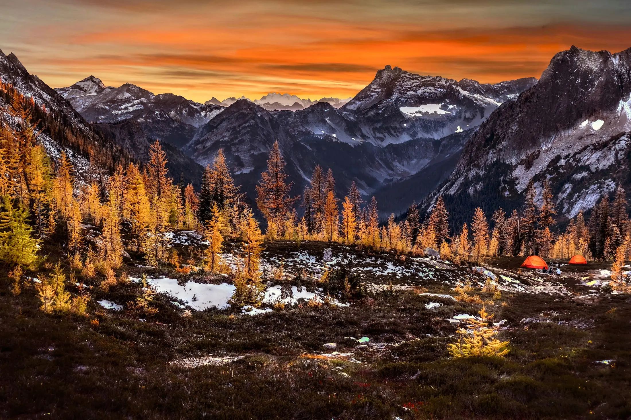 Two orange tents in a larch basin below jagged peaks at sunset